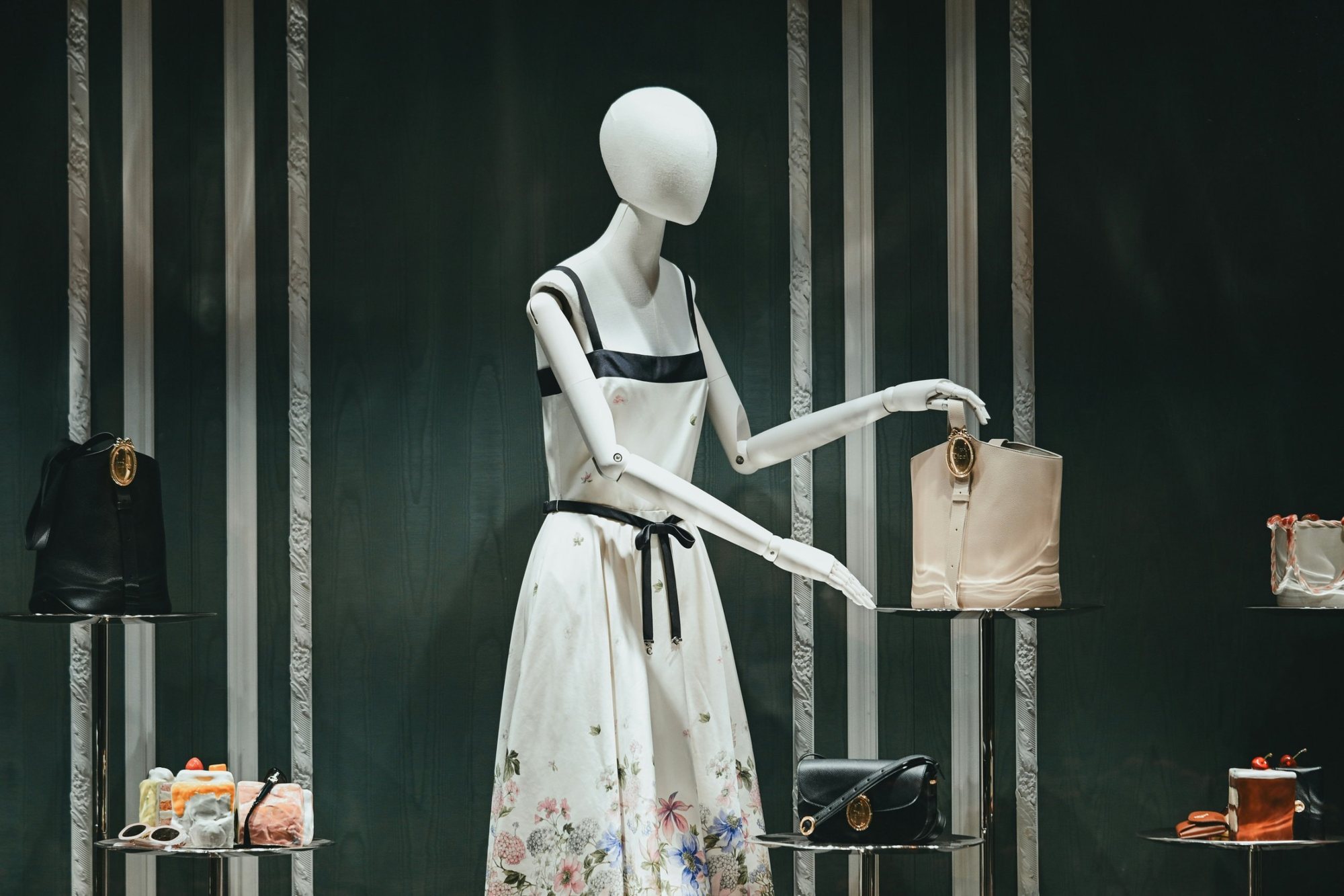 A white mannequin in a floral dress reaching for a cream bucket bag, in a window with dark green moire walls